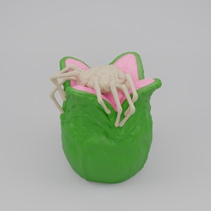 May include: A green, plant-like sculpture with pink detailing, housing a light beige, spider-like creature. The sculpture has a textured surface and a rounded shape, with the spider resting inside the pink-lined opening.