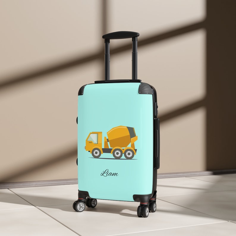 May include: A light blue suitcase with a yellow cement mixer truck graphic and the name "Liam" printed on the front.