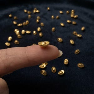 May include: A collection of small, gold-colored metal ingots shaped like traditional Chinese sycee currency. Each piece features a smooth, rounded base and a raised central dome. The ingots are scattered across a dark, textured surface.