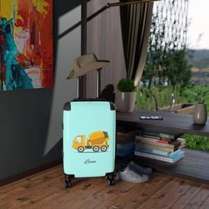 May include: A light blue suitcase with a yellow cement mixer truck design and the name "Liam" printed on the front. The suitcase has a black handle and four black wheels.