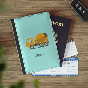 May include: A light blue passport holder with a black border. The front of the holder features a cartoon illustration of a yellow cement mixer truck. The name "Liam" is written in cursive below the truck.