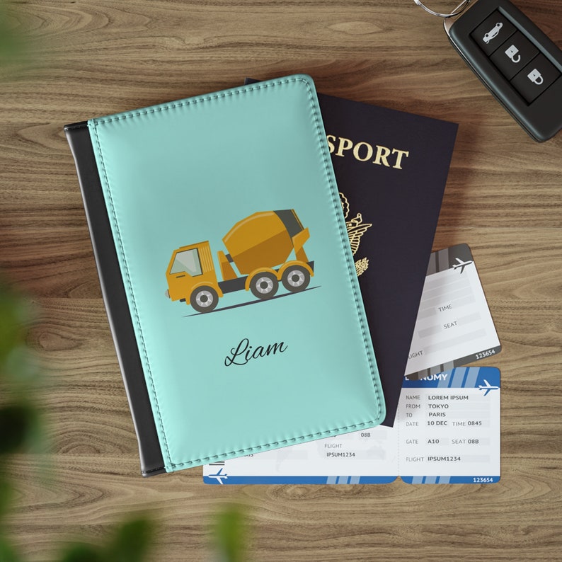 May include: A light blue passport holder with a black border. The front of the holder features a cartoon illustration of a yellow cement mixer truck. The name "Liam" is written in cursive below the truck.