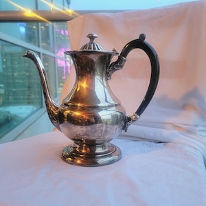 Puede incluir: Una tetera de plata con asa negra. La tetera tiene un cuerpo redondo y un pico en el lateral. La tetera está sobre una superficie blanca.