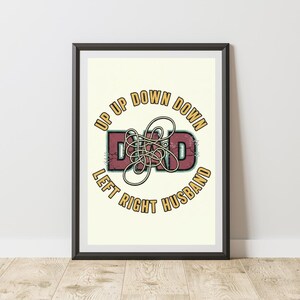 May include: A framed wall art print featuring the text UP UP DOWN DOWN LEFT RIGHT HUSBAND in yellow, arched around a central maroon DAD graphic with a white tangled line overlay. The design has a distressed, vintage-style cracked texture.