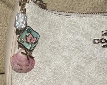 May include: A Coach handbag with a decorative charm. The charm includes a pearl, a teal and pink floral pendant, and a pink seashell. The handbag is white with a repeating pattern and gold hardware.