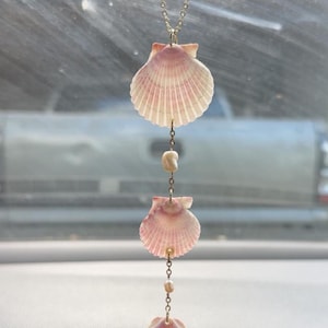 May include: A car charm featuring three pink and white scallop shells suspended by a delicate gold chain. Small, round beads are interspersed between the shells. The charm hangs from a car's rearview mirror.