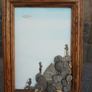 May include: A framed wall art piece featuring a light blue background with a single paper bird cutout. Three figures constructed from small gray stones and thin wooden sticks are positioned on a cluster of larger gray rocks.