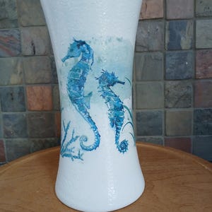 May include: White ceramic vase with a textured surface, featuring blue seahorse illustrations. The vase has a flared top and a rounded base, with a design that evokes an underwater scene. The vase is placed on a wooden surface.