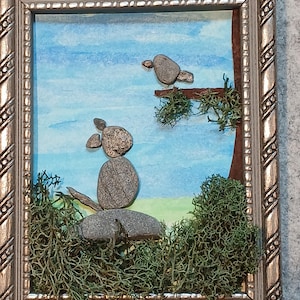 May include: Framed artwork featuring a whimsical scene of birds crafted from smooth, gray stones. The birds perch on a branch and a large stone, set against a watercolor sky and green moss. The silver frame adds a touch of elegance to this nature-inspired piece.