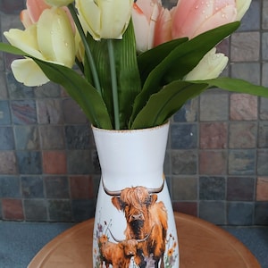 May include: A white vase with a Highland cow design, holding a bouquet of artificial tulips in shades of yellow and pink. The vase has a flared top and a detailed illustration of a cow and calf. The tulips have water droplets.