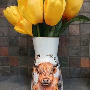 Könnte beinhalten: Eine weiße Vase mit einer Aquarellillustration einer Highland-Kuh und floralen Akzenten, die einen Strauß leuchtend gelber Tulpen enthält. Die Vase hat eine leicht strukturierte Oberfläche und eine ausgestellte Form, was eine rustikale und charmante Ästhetik erzeugt.