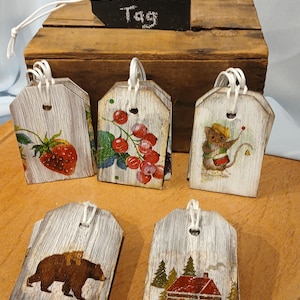 May include: A set of 8 rustic wooden gift tags with various designs, including a bear, a cabin, berries, and a strawberry. The tags are whitewashed and have a distressed look. The tags are tied with white twine and have a chalkboard sign that reads "Reusable Gift Tag".