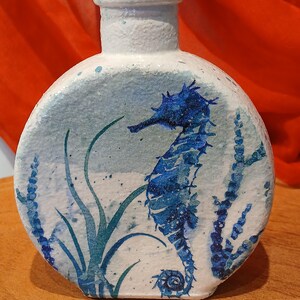 May include: White ceramic vase with a blue and green seahorse design. The vase has a round body and a narrow neck.