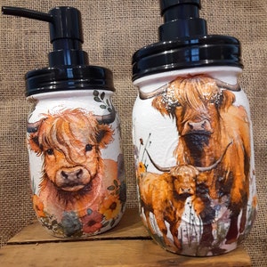 May include: Two glass soap dispensers with black pumps and lids. The dispensers are decorated with a watercolor design of Highland cows and wildflowers.