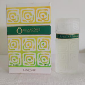 Puede incluir: Un conjunto de perfume con frasco y caja. La caja es blanca con un patrón geométrico amarillo y verde y el texto "Ô DE LANCÔME EAU DE TOILETTE". El frasco es de vidrio esmerilado con una banda verde y el texto "7,5 ml CP 85% vol".