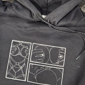 May include: Black hoodie with a white embroidered design of a comic book style image of a woman's body. The image is divided into three panels.