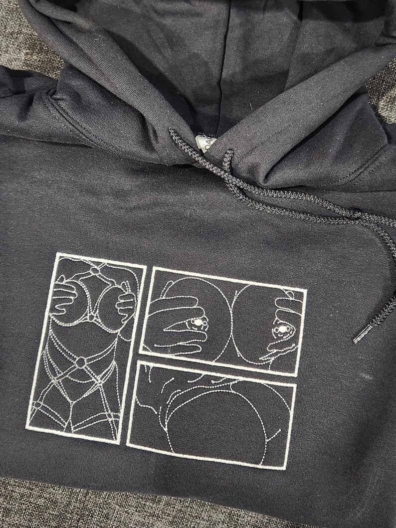 May include: Black hoodie with a white embroidered design of a comic book style image of a woman's body. The image is divided into three panels.
