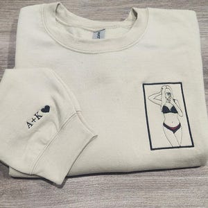 May include: A light beige sweatshirt with a black embroidered image of a woman in a bikini taking a selfie. The sweatshirt has a black embroidered heart and the text "A+K" on the left sleeve.