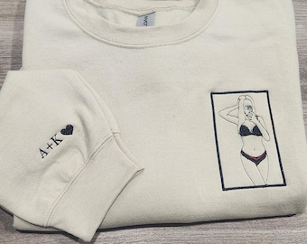 Puede incluir: Una sudadera beige claro con una imagen bordada en negro de una mujer en bikini tomando una selfie. La sudadera tiene un corazón bordado en negro y el texto "A+K" en la manga izquierda.