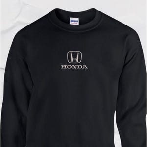 May include: Black crew neck sweatshirt with the Honda logo in silver. The logo features a stylized 'H' within a square. The word "HONDA" is printed below the logo in silver. The sweatshirt is made by Gildan.