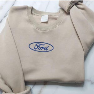 Puede incluir: Sudadera beige de cuello redondo con el logotipo de Ford bordado en azul en el pecho. El logotipo de Ford tiene forma ovalada con la palabra "Ford" en una fuente estilizada. La sudadera tiene cuello y puños acanalados.