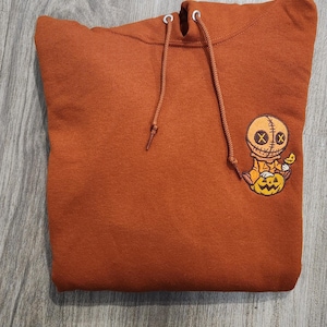 May include: A rust-colored hoodie with a small embroidered image of a stitched-up doll holding a jack-o-lantern.