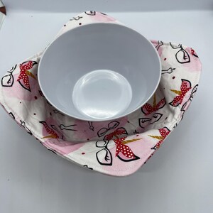 May include: A white ceramic bowl is placed inside a pink and white fabric bowl cosy. The cosy has a repeating pattern of unicorns with red bows and polka dots. The background of the cosy has a watercolour effect.