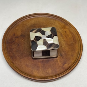May include: A small square box featuring a geometric inlay pattern on the lid, composed of alternating dark brown and iridescent white shell segments. The sides of the box also display a matching pattern of dark brown and white rectangular panels.