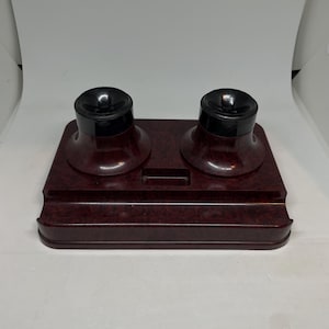 Op de afbeelding: Een vintage bordeauxrode inktpot met twee inktpotten en zwarte deksels. De inktpot is rechthoekig met een gebogen voorkant en een sleuf in het midden. De inktpotten zijn rond en zitten bovenop de inktpot.