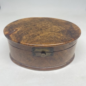 Puede incluir: Una caja de madera de forma ovalada con un patrón de vetas de color marrón moteado. La caja presenta una tapa lisa y redondeada, y un mecanismo de cierre central de metal color latón en la parte frontal, fijado con pequeños tornillos.