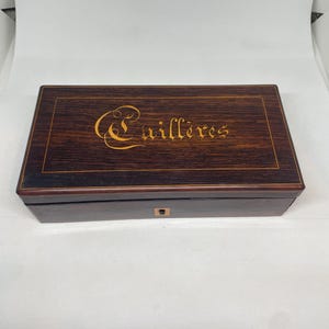 Könnte beinhalten: Eine rechteckige Holzkiste mit dunkelbraunem Finish. Der Deckel trägt den Schriftzug "Cuillères" in goldener Schrift. Die Kiste hat einen helleren Holzrahmen und ein kleines Schlüsselloch auf der Vorderseite.