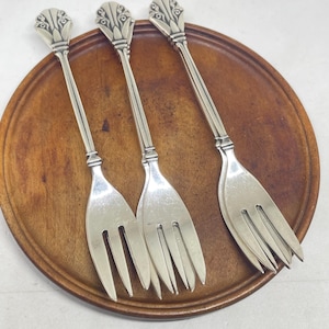 Puede incluir: Tres tenedores de metal plateado con mangos largos y delgados y remates decorativos en forma de abanico. Cada tenedor tiene tres dientes y una superficie pulida y reflectante. Los mangos son rectos con una sutil cresta vertical central.