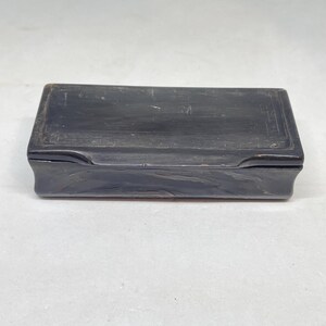 May include: A rectangular black box with a smooth, dark finish and a hinged lid. The surface features subtle, worn textures and faint, etched linear detailing along the edges of the top cover, presenting a simple, functional storage container.