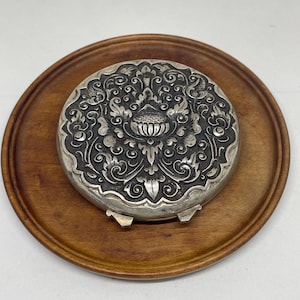 May include: A silver compact with an intricate floral design, resting on a wooden tray.