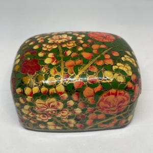 Op de afbeelding: Een decoratieve, handbeschilderde doos met een bloemenontwerp. De doos is voornamelijk groen met rode, gele en oranje bloemen. Gouden accenten benadrukken het bloemmotief. De doos heeft een afgeronde rechthoekige vorm.