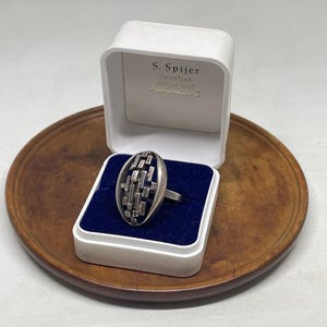 Puede incluir: Un anillo de plata con una cara ovalada que presenta una rejilla de barras rectangulares en relieve, presentado en un joyero blanco con interior de terciopelo azul. La tapa muestra el texto S. Spijer Juwelier, Koningsplein 1, Amsterdam-C.