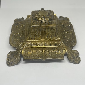 May include: An antique brass inkwell, featuring a square ink pot at its centre, adorned with floral and scrollwork. The inkwell has a rectangular base with curved, leaf-shaped extensions. The brass has a warm, aged patina, showing its age.