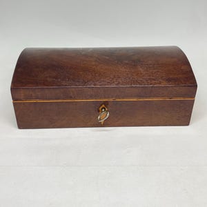 Puede incluir: Una caja rectangular de madera con acabado marrón oscuro y tapa arqueada. La caja presenta una incrustación de color claro alrededor de la costura central y un escudo de metal para la cerradura con una pequeña llave plateada insertada.