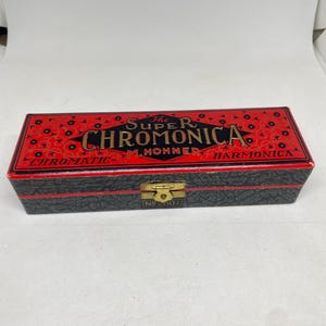 May include: A vintage black box with red detailing, for a Super Chromonica M. Hohner chromatic harmonica. The box features gold lettering that reads "The Super Chromonica M. Hohner Chromatic Harmonica".