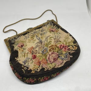 Könnte beinhalten: Eine antike Gobelin-Handtasche mit floralem Design. Die Tasche hat einen beigefarbenen Hintergrund mit bunten Blumen in Rosa, Rot, Blau und Gelb. Die Ränder sind dunkelbraun mit Perlenbesatz und sie hat einen goldfarbenen Kettenriemen.