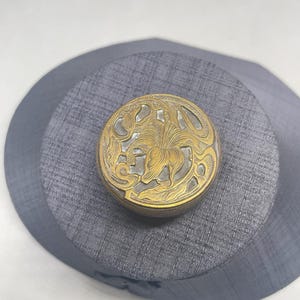 May include: A circular metal container featuring an intricate gold-toned relief design of a central flower surrounded by swirling, organic patterns. The lid has a textured, dark metallic background that contrasts with the raised, polished gold-colored floral motif.