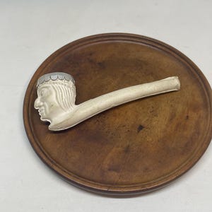 May include: A white pipe with a Native American profile on the bowl, resting on a round, wooden, brown tray. The pipe has a long, straight stem. The bowl features a detailed face and headdress.