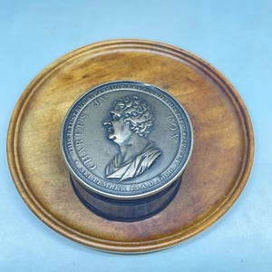 Puede incluir: Un medallón circular de tono bronce con un perfil en relieve de una persona, con la inscripción "CHARLES JAS FOX" y "THIS ILLUSTRIOUS PATRIOT DEPARTED THIS LIFE SEPTEMBER 13 A.D. 1806 AET 57", montado sobre una base de madera oscura.