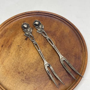 May include: Two antique silver serving forks with rose-shaped handles and three tines each, resting on a round wooden tray. The forks have intricate floral detailing and a vintage aesthetic, perfect for serving appetizers or desserts.