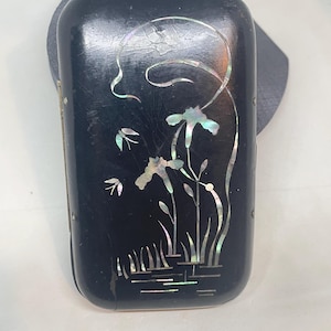 May include: A black rectangular box with a floral design inlaid with iridescent mother-of-pearl. The design features stylized flowers, leaves, and water elements. The box has a slightly rounded top and is set against a gray background.
