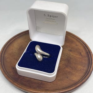Può includere: Un anello bypass in argento dal design liscio e organico a forma di goccia, posizionato all'interno di un portagioie bianco con interno in velluto blu scuro. Il coperchio riporta il testo stampato S. Spijer Juwelier, Koningsplein 1, Amsterdam-C.