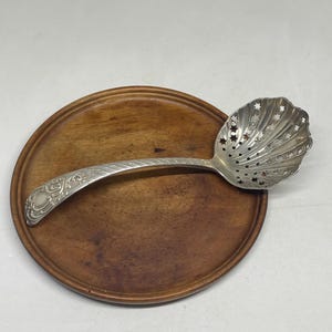 Op de afbeelding: Een zilverkleurige zeeflepel met een schelpvormige bak voorzien van stervormige perforaties en een gebogen handvat met in reliëf aangebrachte krulpatronen langs de steel en op het afgeronde uiteinde van het handvat.