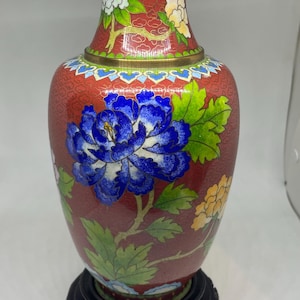 Pode incluir: Um vaso cloisonné vermelho com padrões florais vibrantes em azul, branco e amarelo com folhas verdes. O vaso tem uma borda de latão no topo e na base, e repousa sobre um suporte de madeira escuro e circular com detalhes esculpidos.