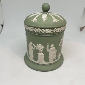 May include: A sage green ceramic jar with a lid and a small spherical knob. The jar features white relief designs of classical figures and floral patterns. The jar is a decorative storage container.