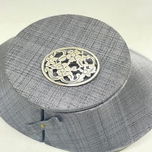 Può includere: Una spilla ovale in argento caratterizzata da un intricato design traforato con fiori e foglie intrecciate. Il metallo presenta una finitura lucida con aree incavate che mettono in risalto i dettagli floreali su tutta la superficie.